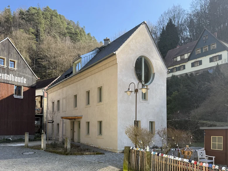 Ein schmales hohes hell verputztes Haus mit einem markanten großen runden Fenster im Gie-bel, daneben und dahinter je ein Haus, im Hintergrund erhebt sich ein bewaldeter Berghang, im Vordergrund ein Lattenzaun mit bunten Tassen auf jeder einzelnen Latte