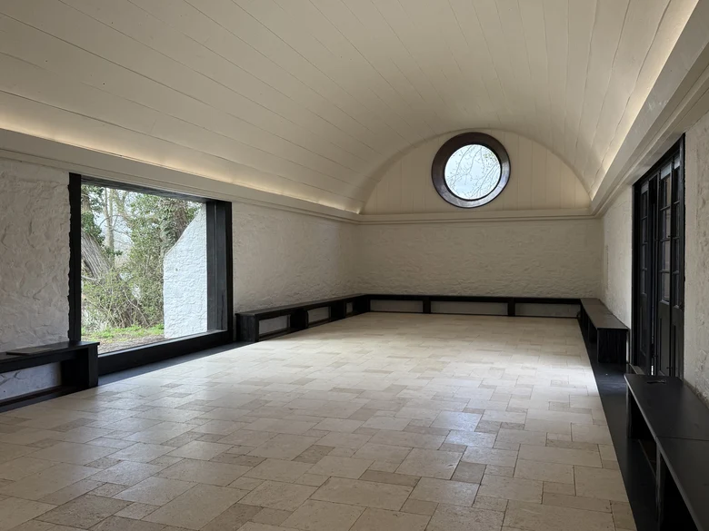 das Innere einer Scheune mit einem Tonnengewölbe aus weißen Holzbrettern, der Blick wird auf ein großes rundes Fenster auf der Giebelseite gelenkt, in der linken Wand ist ein großes bodentiefes Fenster zu sehen, der Fußboden besteht aus hellen Fliesen, an