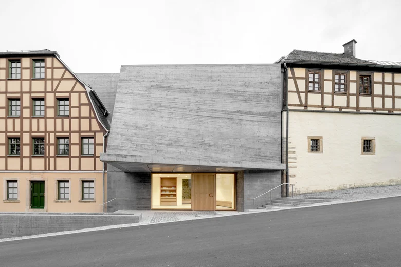 Ein modernes Gebäude mit geschwungenem Betondach steht zwischen zwei Fachwerkhäu-sern an einer nach links abschüssigen Asphaltstraße, der Eingangsbereich ist hell erleuchtet