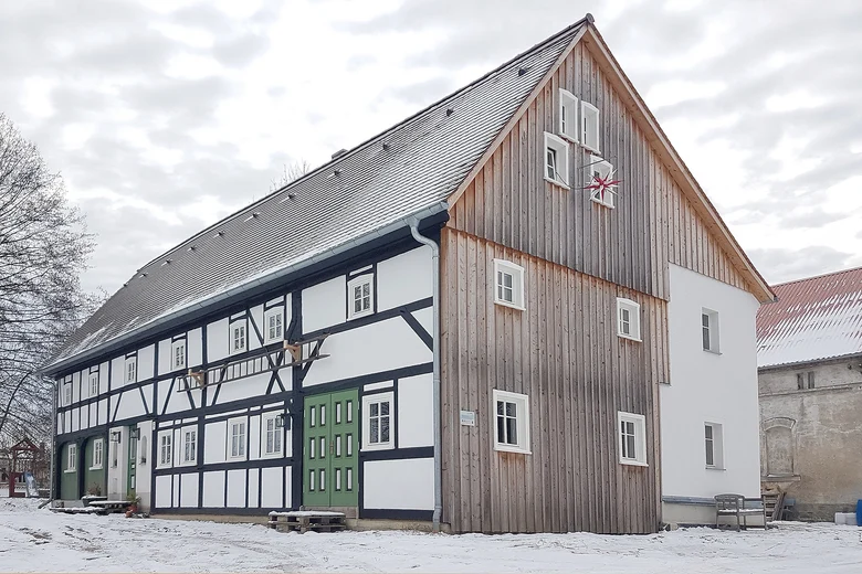 Ein großes saniertes Umgebindehaus, man sieht den hohen mit Holz verschalten Giebel, an dem außen ein rot-weißer Herrnhuter Stern hängt und die Längsseite mit dunklen Balken und weiß verputzten Gefachen, der Himmel ist bewölkt, rings um das Haus liegt ein