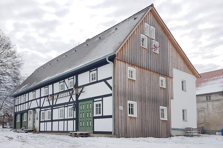 Ein großes saniertes Umgebindehaus, man sieht den hohen mit Holz verschalten Giebel, an dem außen ein rot-weißer Herrnhuter Stern hängt und die Längsseite mit dunklen Balken und weiß verputzten Gefachen, der Himmel ist bewölkt, rings um das Haus liegt ein